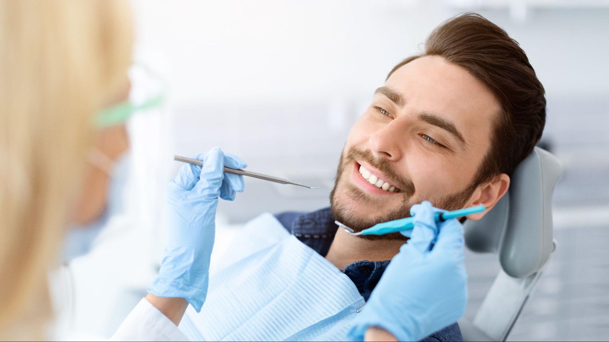 Female dentist holding tools next to a smiling man in a dental exam chair.