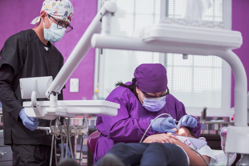 A female dentist performs dental treatment on a young girl while the male assistant observes.
