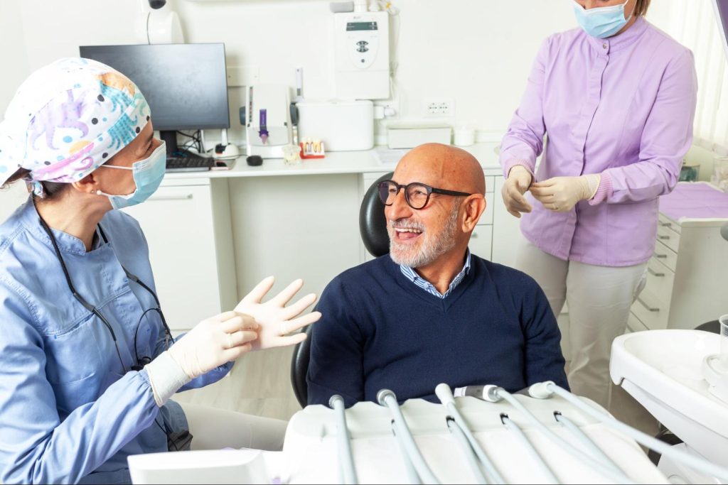 A lady dentist wearing gloves talks with a senior male patient during a denture consultation.