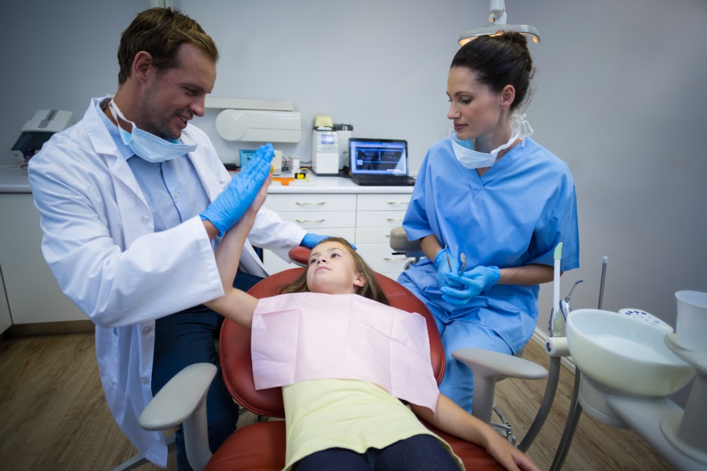 A little girl is lying on the dentist chair, high-fiving a male dentist.
