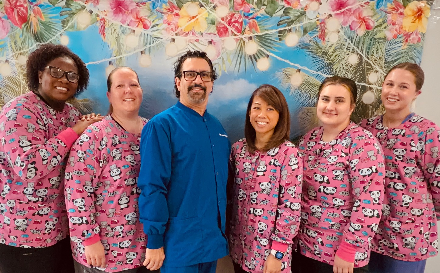 Six staff members in pink panda-print scrubs pose with a man in blue scrubs in front of a tropical floral backdrop, smiling.
